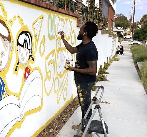 Tysen Knight paints portion of long mural on stucco wall, lining a city sidewalk, depicting students reading a book