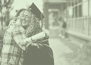 Female high school graduate wearing gown and holding diploma smiles as she hugs a person wearing a plaid shirt