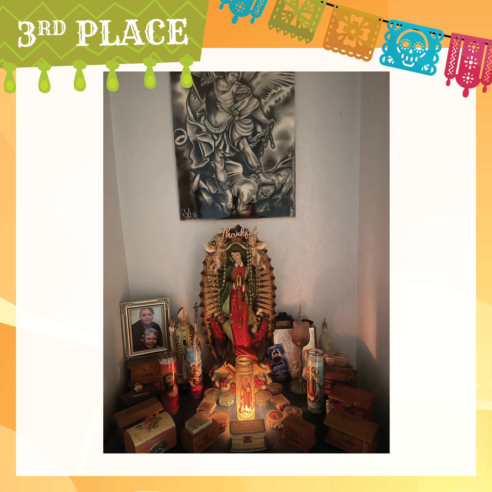 photo graph of dia de los muertos alter, with graphite drawing of male angel holding down man with his foot and the alter below with a figure of Mary, holy candles, a figure of the pope, a photo of two women and an array of small boxes