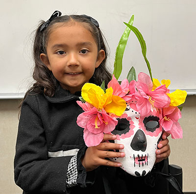 Olivia holding her Dia de los muertos white mask with pink accents and pink and yellow flowers