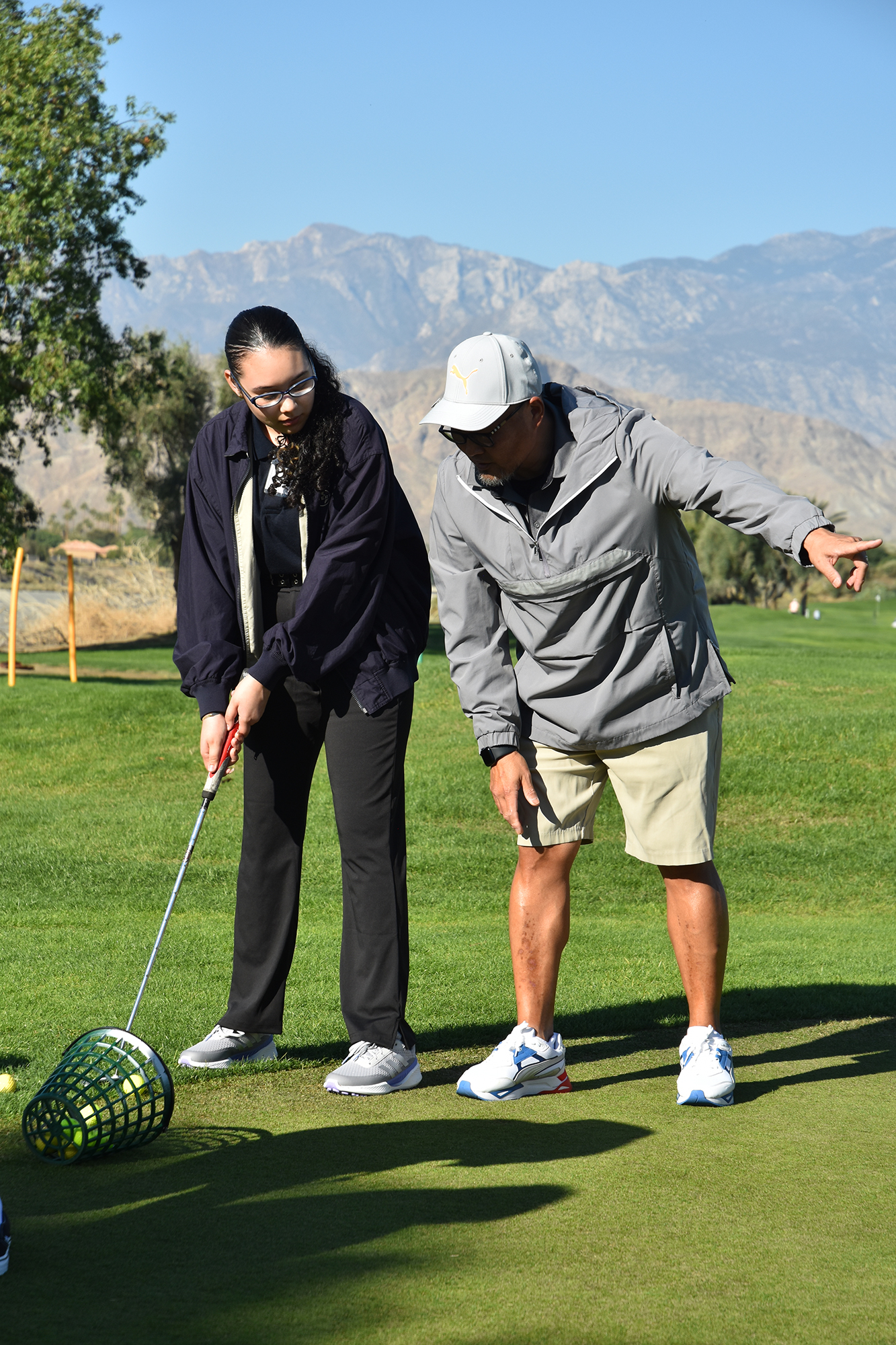 Yuritza stands on a golf course next to coach mimicking his stance while aligning golf club for a swing