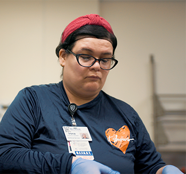 Young woman wearing hair net, medical gloves, and multiple hospital employment badges concentrating on her work