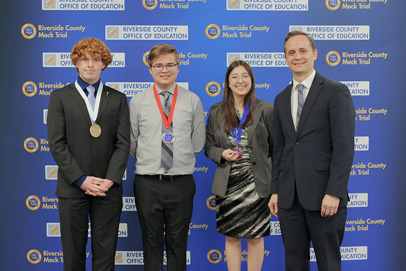 Three student honorees pose and lawyer in front of Riverside County Mock Trial backdrop