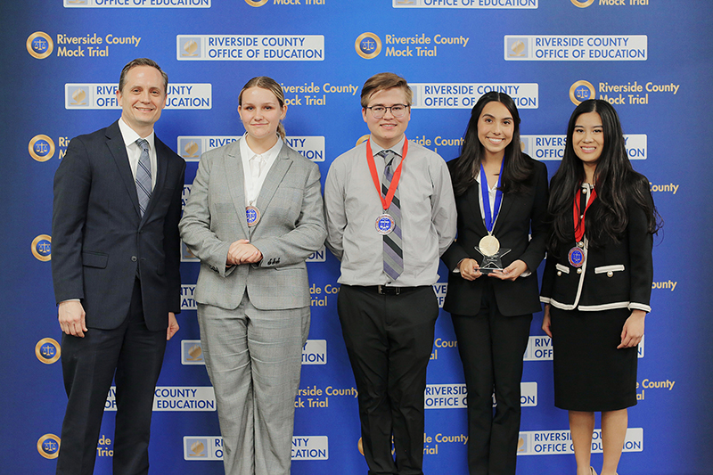 Four student honorees pose with lawyer in front of Riverside County Mock Trial backdrop