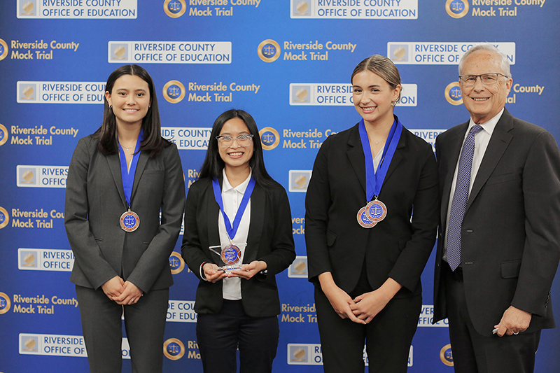 Three student honorees pose with lawyer in front of Riverside County Mock Trial backdrop